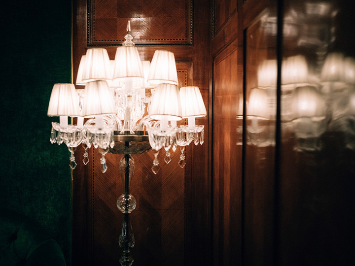 Elegant crystal chandelier with small lampshades, which stands in front of a wood-paneled wall and creates a warm, stylish atmosphere.
