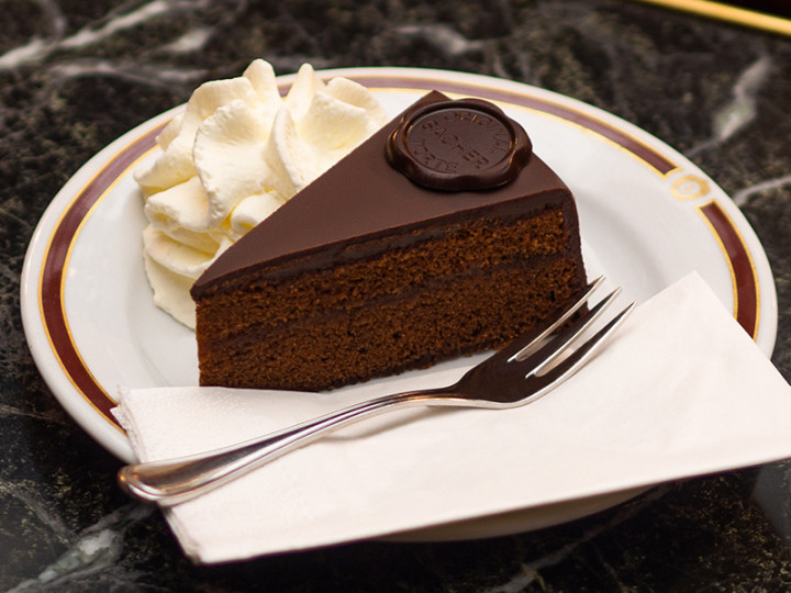 Piece of original Sacher cake with whipped cream on a porcelain plate, served with a fork and napkin.
